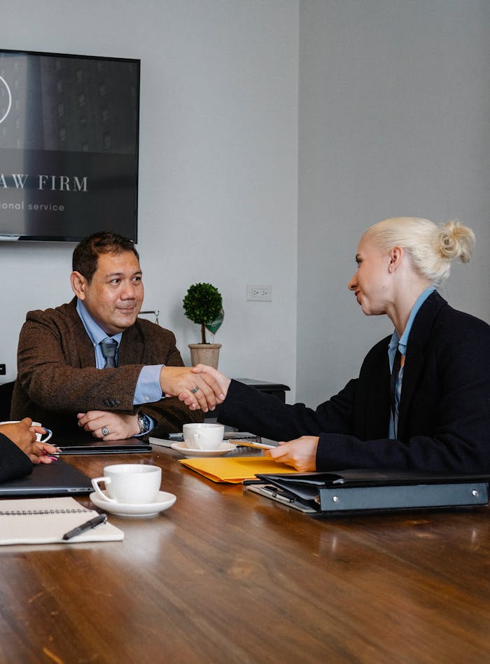 about-us Smiling multiracial coworkers in formal clothes gathering at wooden table with cups of coffee and various folders and shaking hands while looking at each other